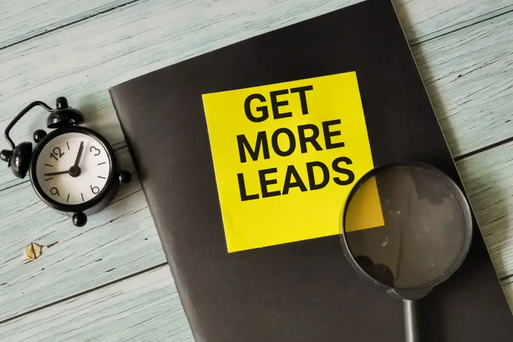 Wooden table top with a black traditional alarm clock with bells, a document with a black cover with get more leads in black caps on a yellow square on the cover and a magnifying glass on top.