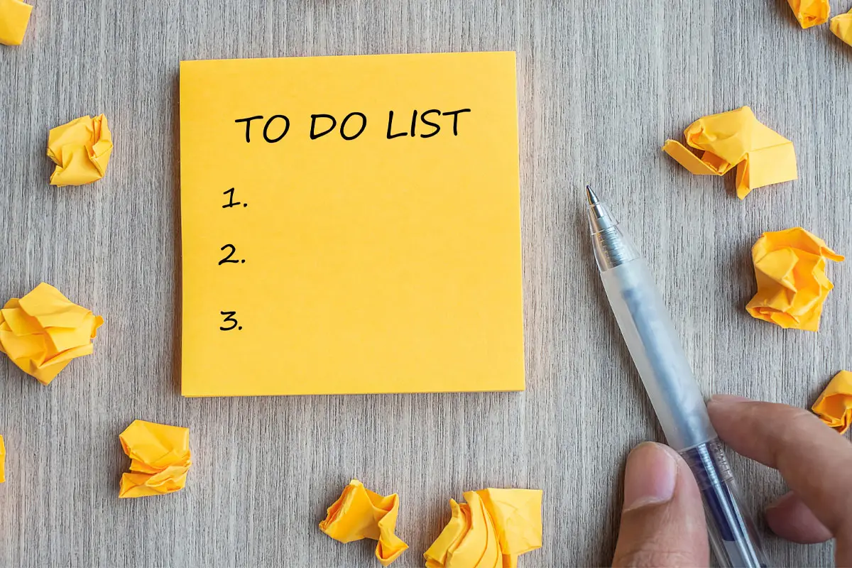 Yellow Square Of Paper With To Do List And Numbers 1, 2 And 3 Written On It. Two Fingers Hold A Pen To The Right Of The Note And There Are Balls Of Yellow Paper Dotted Around The Surface.